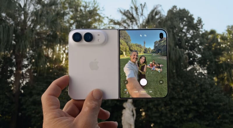 A hand holds a white foldable smartphone, displaying a camera interface on its screen that shows a man and woman taking a selfie in a park.