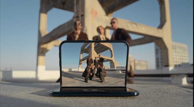 A foldable smartphone sits on a concrete ledge, displaying a camera viewfinder with face-tracking boxes over a group of five people posing in front of a large concrete structure.