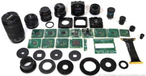 A collection of disassembled camera components, including lenses, circuit boards, sensor modules, and various mounting rings, laid out on a white surface.
