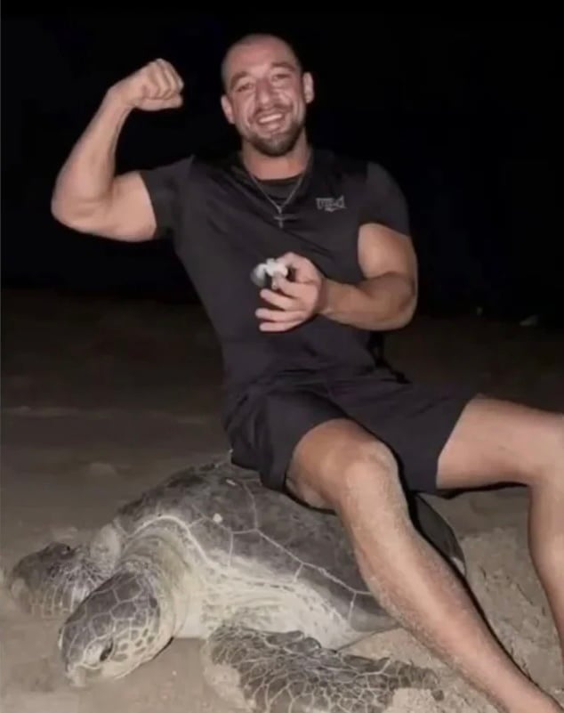 A man smiling and flexing his arm while sitting on the back of a large sea turtle on a beach at night.