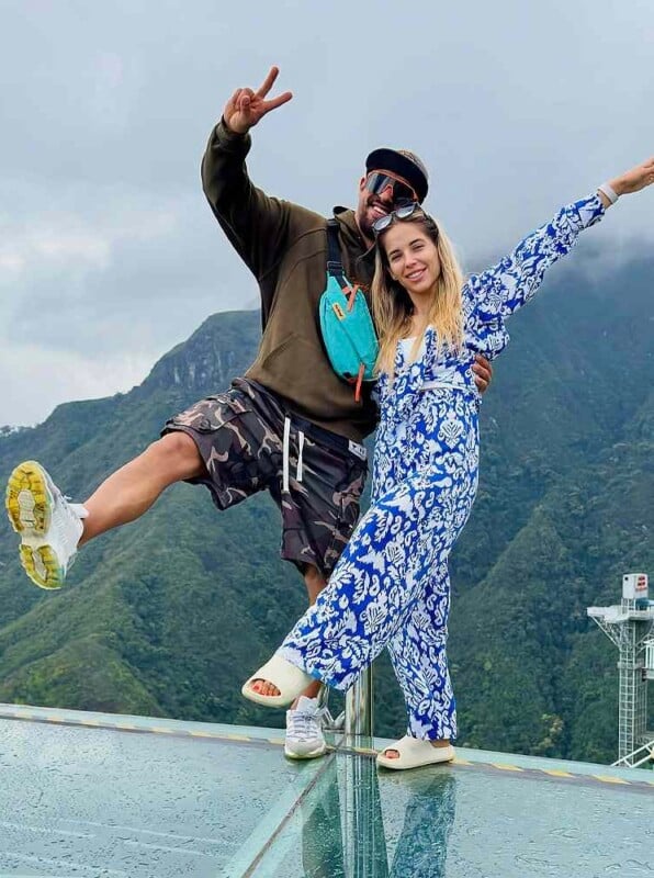 A man and a woman pose on a glass observation deck overlooking a mountain range, with the man making a peace sign and the woman waving.