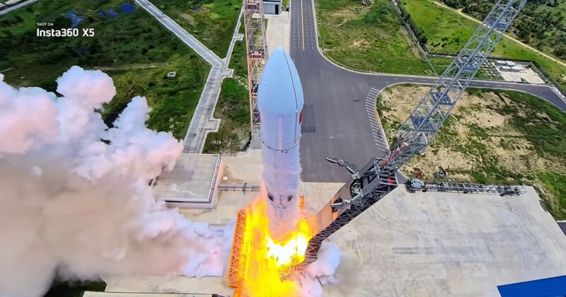A rocket launches from a pad, surrounded by bright orange flames and large plumes of white smoke, with a tall service tower visible to the right.