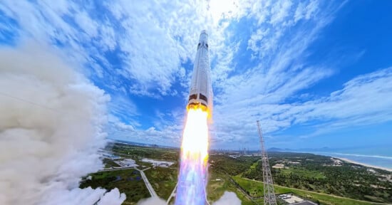 A rocket launches into a bright blue sky, leaving a large plume of white smoke over a coastal landscape.