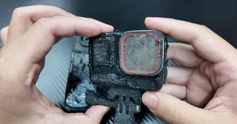 A pair of hands holds a heavily mud-caked action camera with a red-rimmed lens cover.