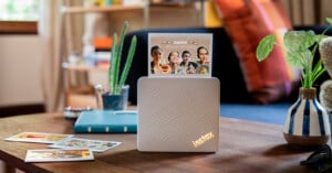 A white Instax printer sits on a wooden table with a printed photo displaying "Happy Anniversary" standing upright in its slot, surrounded by other photos, a small cactus, and a potted plant.