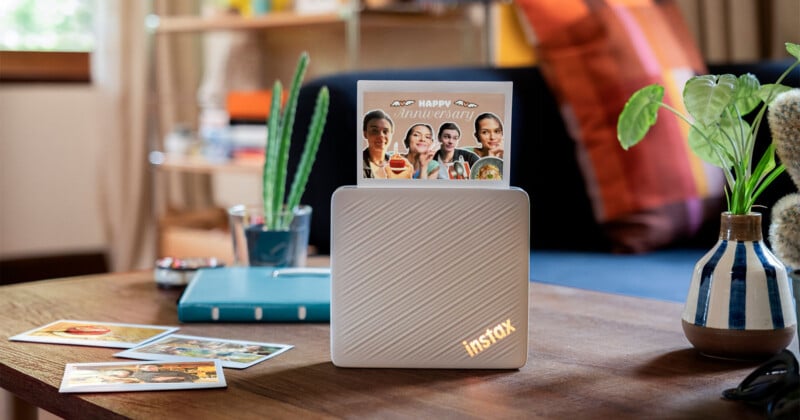 A white Instax printer sits on a wooden table with a printed photo displaying "Happy Anniversary" standing upright in its slot, surrounded by other photos, a small cactus, and a potted plant.