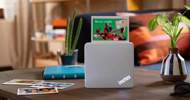 A white Instax printer sits on a wooden table next to a potted plant, a blue notebook, and several printed photos, with one photo emerging from the top displaying a "Movie night" graphic.