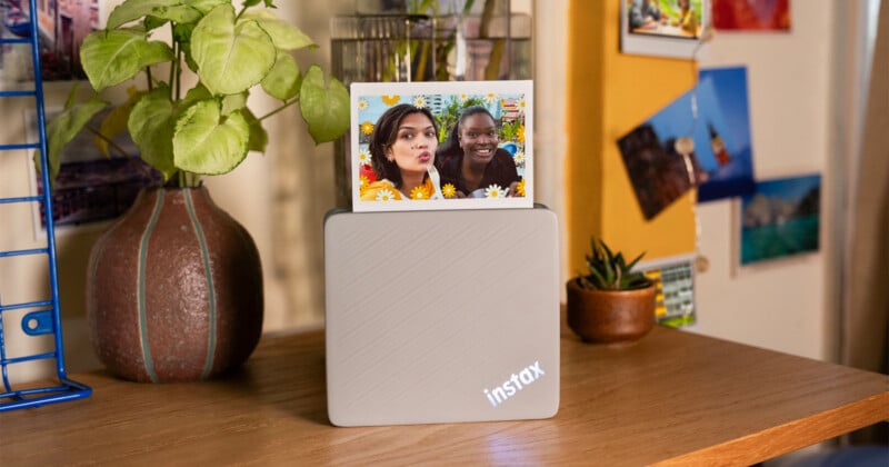A light gray Instax printer sits on a wooden desk with a small photo of two women emerging from the top, next to a potted plant and a blue wire rack.