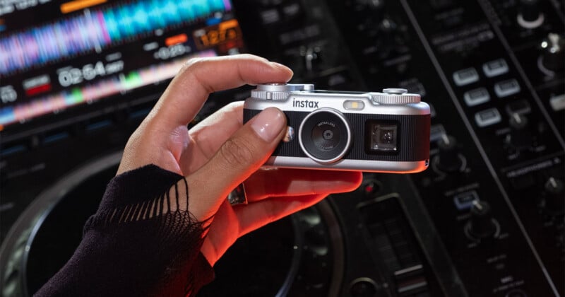 A hand with a black sleeve holds a silver and black Instax camera in front of a DJ mixing console.