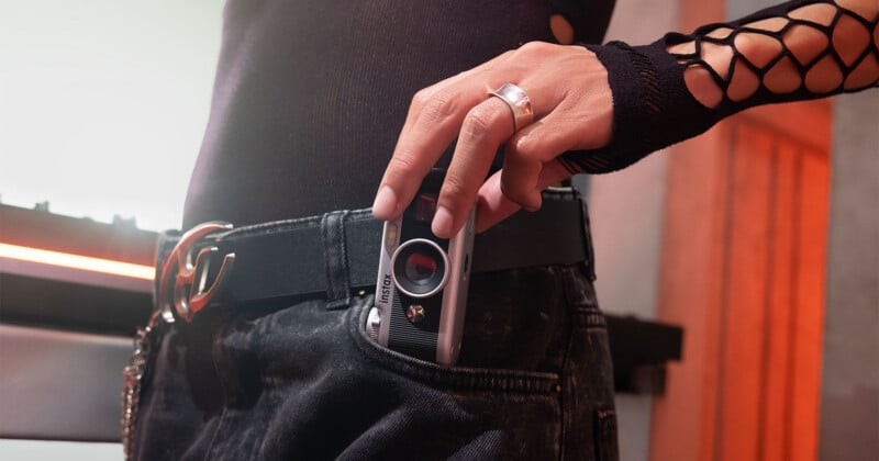 A person wearing a black top and jeans with a fishnet sleeve holds a small silver camera partially inside their pocket.