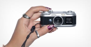A hand with metallic pink nails and silver rings holds a small, silver and black Instax digital camera against a white background.