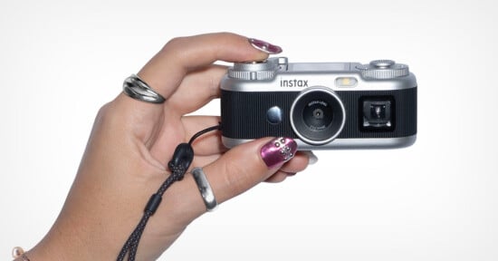 A hand with metallic pink nails and silver rings holds a small, silver and black Instax digital camera against a white background.