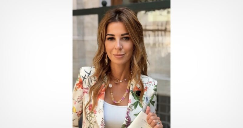 A woman with long, wavy brown hair wears a white floral blazer over a white top, layered necklaces, and dangling earrings while holding a small white booklet.