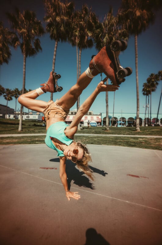 A woman in a turquoise top and tan shorts performs a one-handed handstand on a paved surface while wearing roller skates.