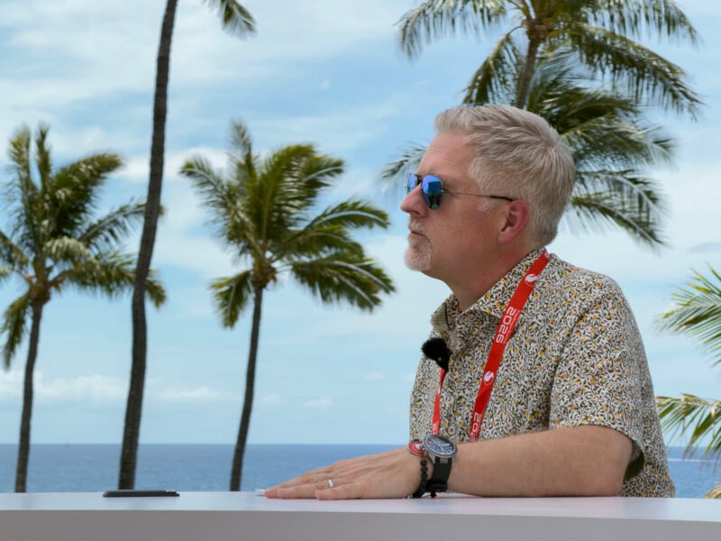 A man with gray hair and sunglasses sits at a table outdoors with palm trees and the ocean in the background, wearing a patterned shirt and a red lanyard.