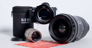 A camera body with a Zeiss lens attached sits behind a smaller silver lens, a large black zoom lens, a KEH Camera branded case, and a red cleaning cloth.
