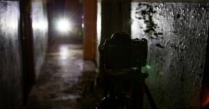 A camera on a tripod faces a bright light source at the end of a dark, narrow corridor with textured walls.