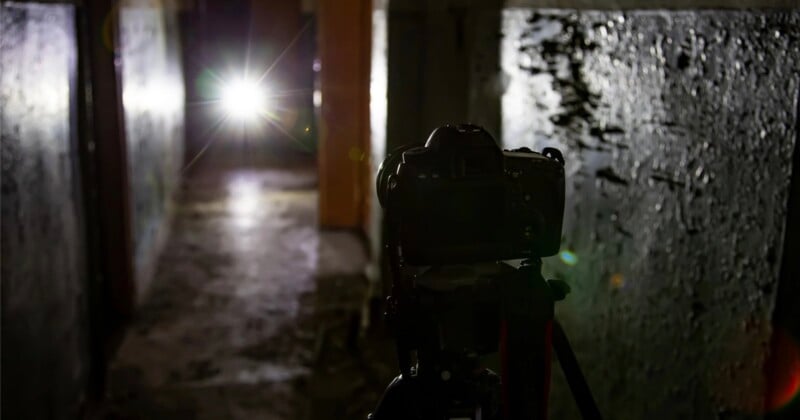 A camera on a tripod faces a bright light source at the end of a dark, narrow corridor with textured walls.