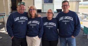 Four people stand side-by-side in an airport terminal wearing matching navy blue sweatshirts that say "LAKE AMERICA" with a wave and anchor graphic.