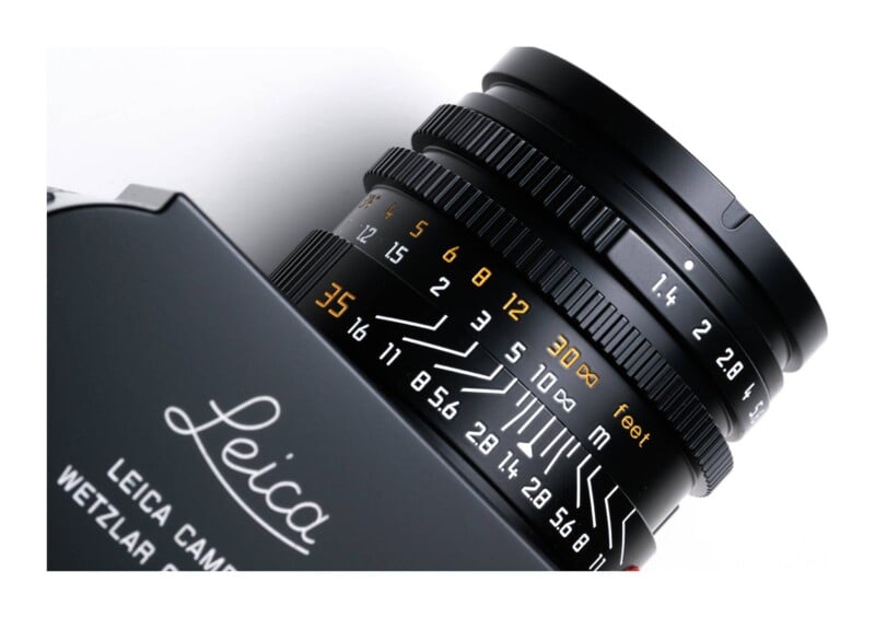 A close-up view of a black Leica camera lens showing the aperture ring, focus scale in feet and meters, and depth of field markings.