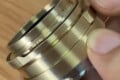 A person holds a small, cylindrical brass camera lens adapter between their thumb and fingers.