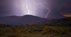 Two lightning bolts strike the peaks of dark, rolling mountains above a grassy field at twilight.