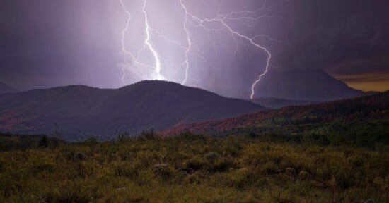 Two lightning bolts strike the peaks of dark, rolling mountains above a grassy field at twilight.