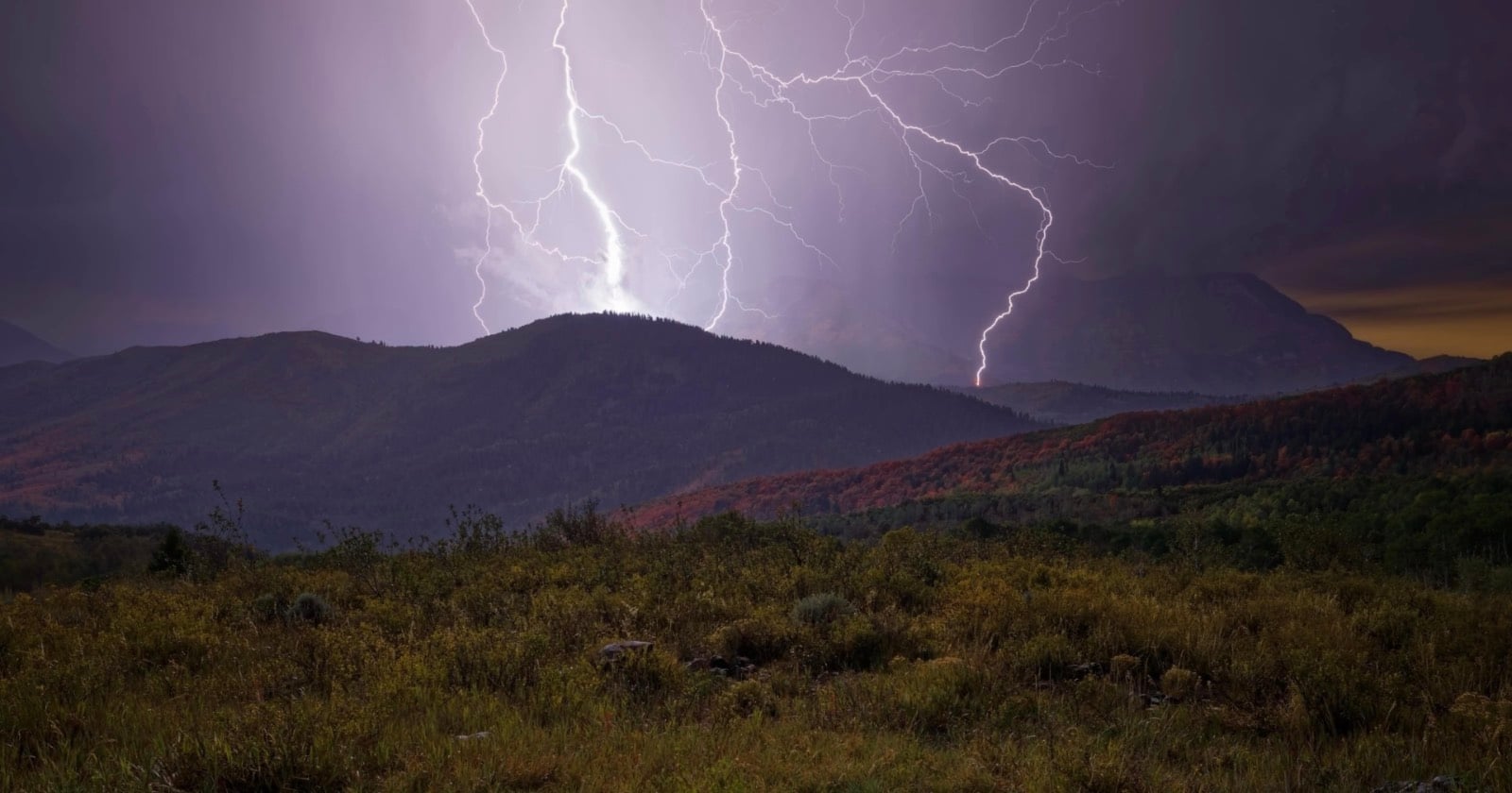 Two lightning bolts strike the peaks of dark, rolling mountains above a grassy field at twilight.