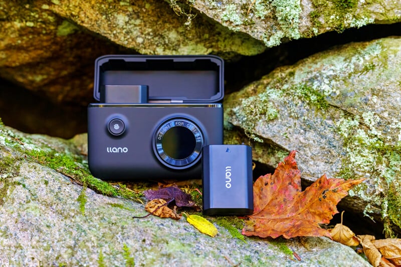 A black Llano charging case with its lid open sits on a mossy rock next to a matching battery pack and a fallen autumn leaf.