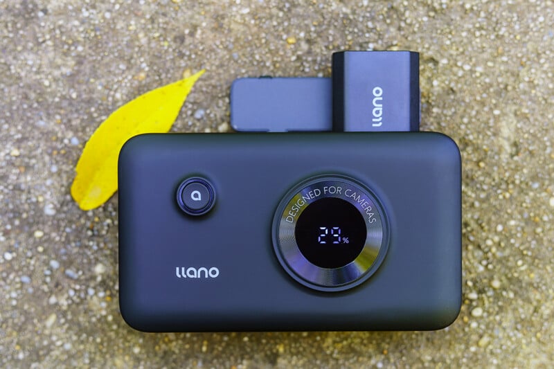 A black Llano portable power bank with a digital display showing 29% battery sits on a textured ground surface next to a yellow leaf.