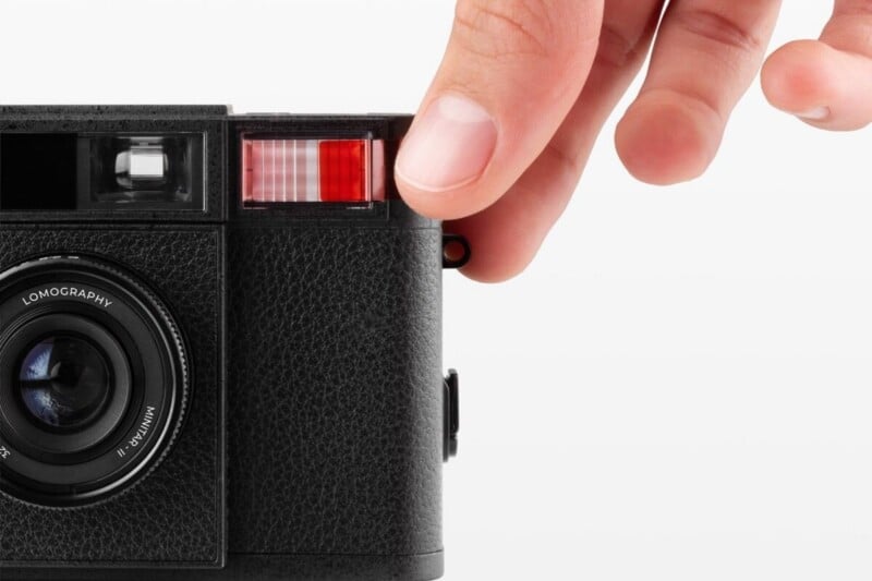 A person's hand presses down on the top corner of a black Lomography Minitar-II camera against a white background.
