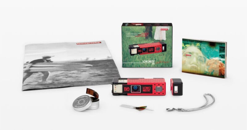 A Lomomatic 110 camera kit including the camera, a detachable flash, a film canister, a strap, a photo book, and a folded poster on a white background.