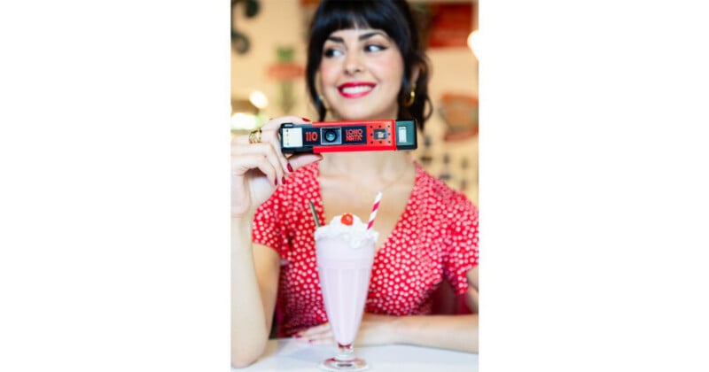 A woman with dark hair and red lipstick holds a red LomoMatic 110 camera in front of her face, with a milkshake in the foreground.