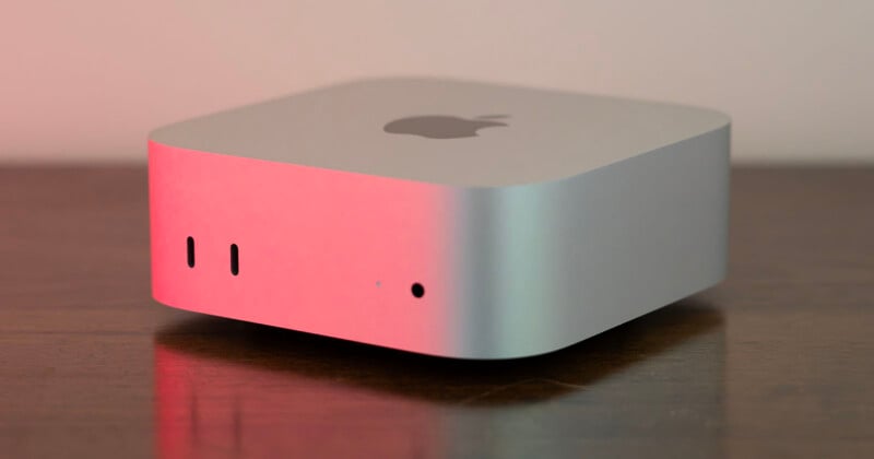 A silver, square-shaped computer with rounded corners sits on a wooden surface, illuminated by red and green light.