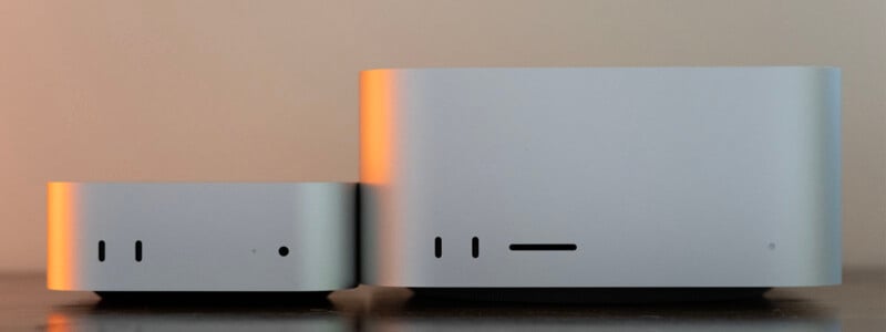 Two silver, box-shaped computer devices sit side-by-side on a dark surface, with the device on the right being significantly taller than the one on the left.