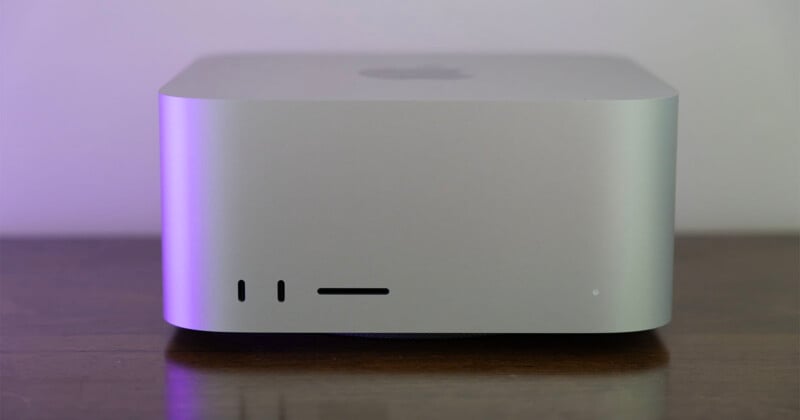 A silver Apple Mac Studio computer sits on a wooden surface, showing two front-facing USB-C ports, an SD card slot, and a status light.
