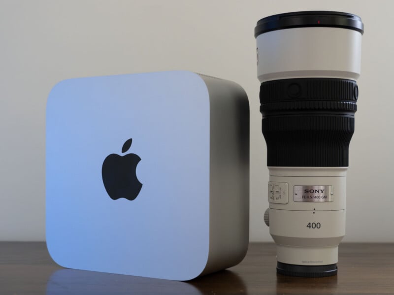 An Apple Mac Studio computer sits next to a Sony FE 4.5/400 GM camera lens on a wooden surface against a plain wall.