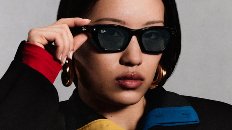 A person with dark hair wears black Ray-Ban sunglasses and large gold hoop earrings, with their hand resting on the frame.