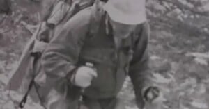 A person wearing a bucket hat and a heavy backpack hikes uphill while using trekking poles.