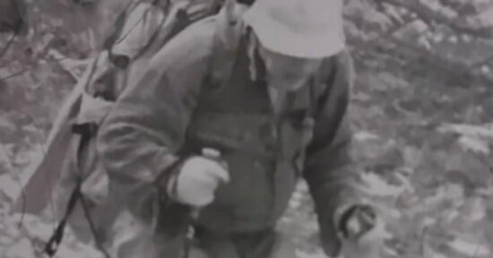 A person wearing a bucket hat and a heavy backpack hikes uphill while using trekking poles.