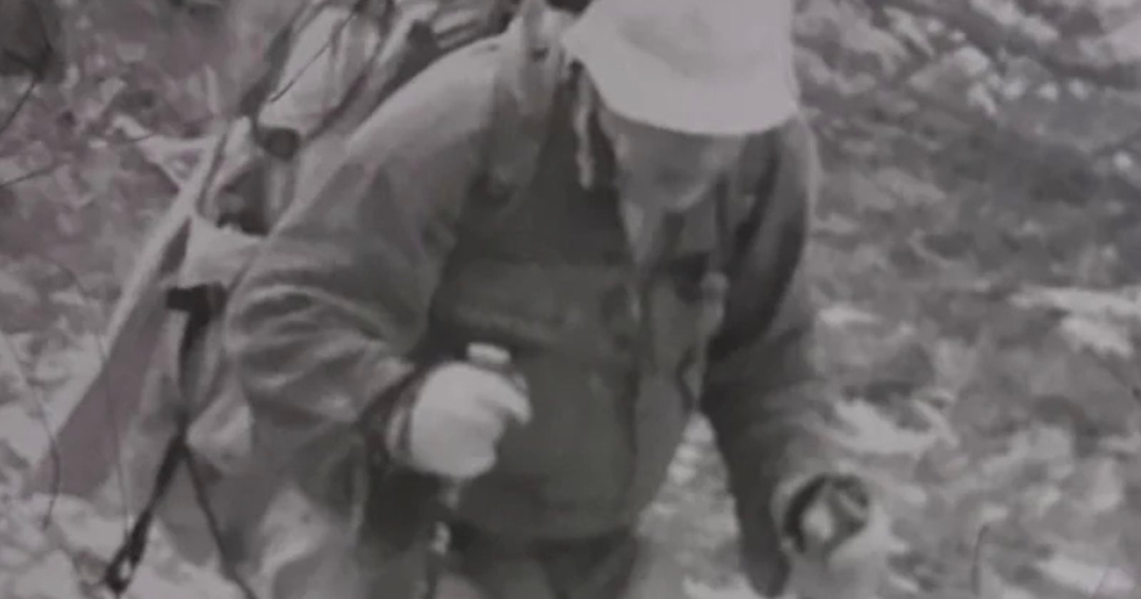A person wearing a bucket hat and a heavy backpack hikes uphill while using trekking poles.
