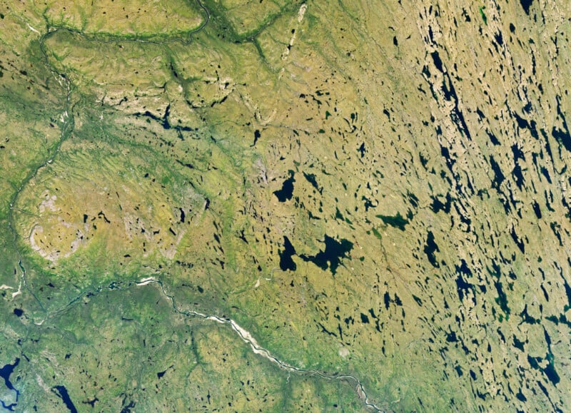 An aerial view of a vast, rugged landscape featuring a winding river, numerous scattered lakes, and patches of green vegetation.