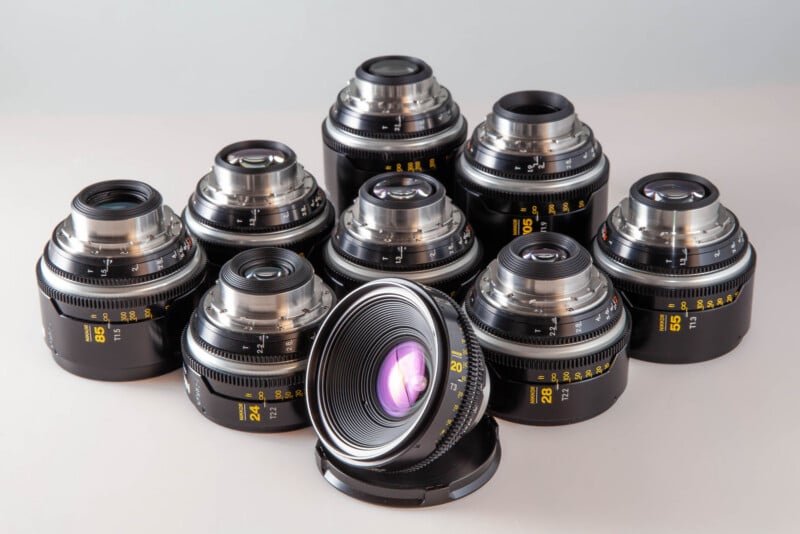 A collection of nine black cinema lenses arranged on a light surface, with one lens positioned in the foreground facing forward.