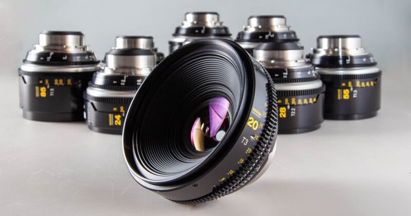 A collection of seven black cinema lenses arranged on a light surface, with a 20mm lens in the foreground.