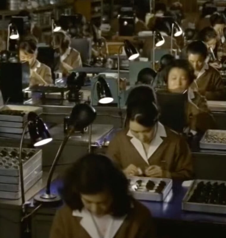 A high-angle shot shows rows of women in brown uniforms working at desks with individual lamps, assembling small components in trays.