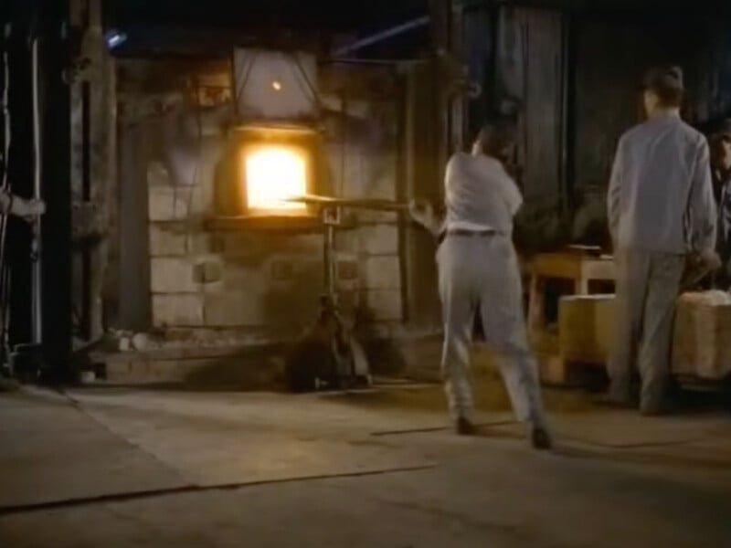A worker in light-colored clothing uses a long metal tool to reach into a glowing industrial furnace while another person stands nearby.