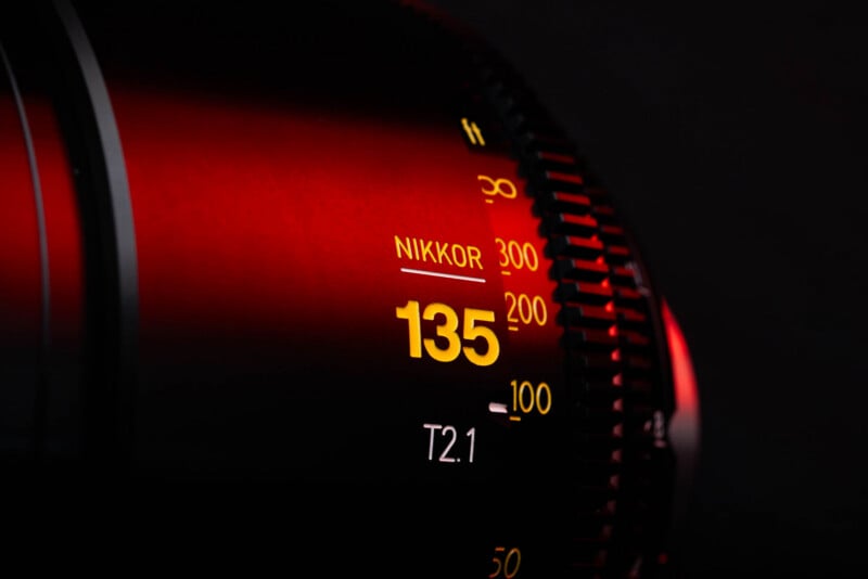 A close-up of a camera lens barrel featuring yellow text that reads "NIKKOR", "135", and "T2.1" against a dark, red-lit background.