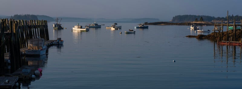 A calm harbor at dusk features several fishing boats moored near wooden piers and rocky shorelines under a hazy sky.