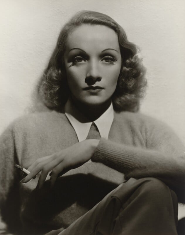 A black-and-white portrait of Marlene Dietrich wearing a collared shirt and sweater, holding a cigarette in her right hand.
