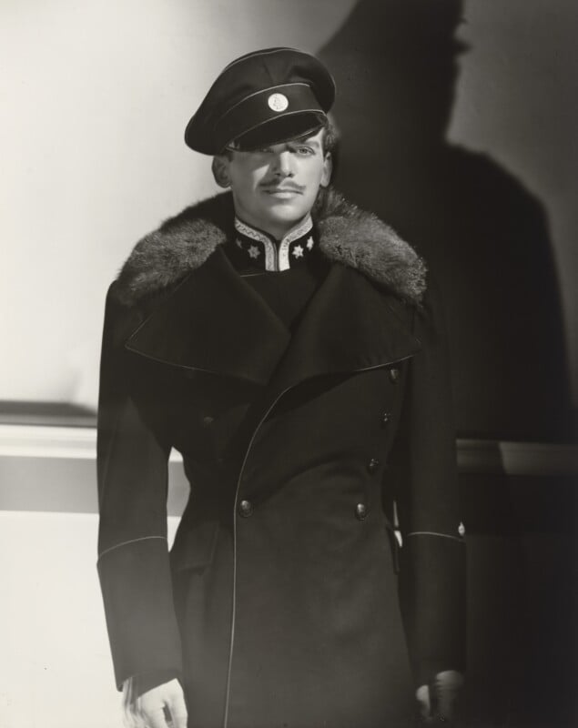 A man with a thin mustache wears a dark, fur-collared military-style coat and a peaked cap with a circular emblem.